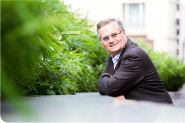Smiling man wearing sport coat leaning against ledge and looking off into the distance.