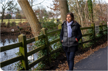 Woman wearing winter coat and scarf, walking on trail alongside a pond.