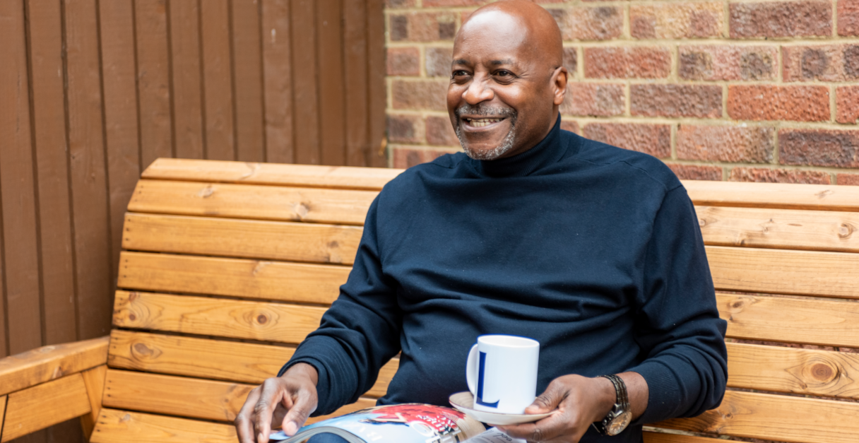 Man sitting on a bench, reading a magazine, and holding a cup of coffee, looking forward and smiling at something out of frame.