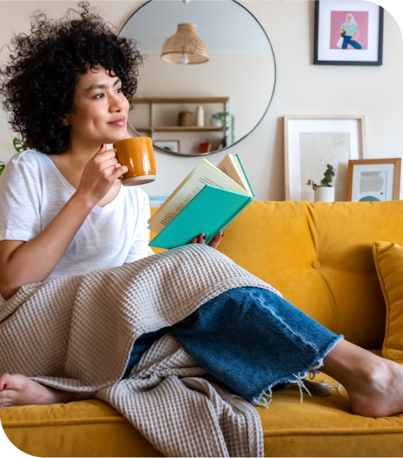 Smiling lady holding a mug and reading a book while sitting on a couch with a blanket over her lap