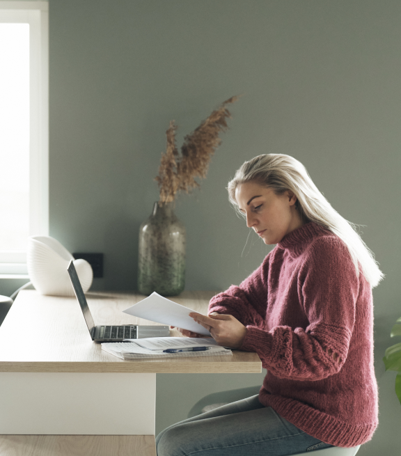 Young girl sitting at a desk with an open laptop and notebook in front of her, holding and reading from a piece of paper