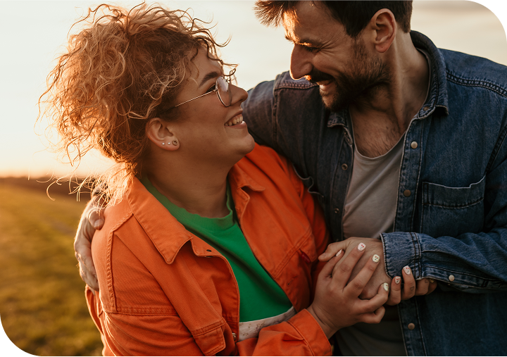 A man and woman hug while looking at each other and smiling with a sunset in the background